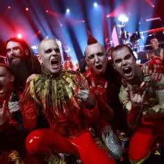 Zespół z Eurowizji Wielka Piątka w czerwonych strojach z błyszczącymi elementami, krzyczący i pozujący na scenie.