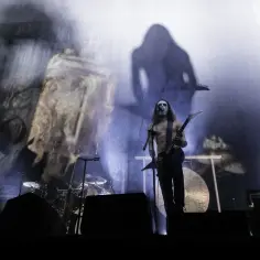 Mroczna scena z wokalistą w makijażu, grającym na gitarze. W tle projekcje i perkusja, tworzące atmosferę **heavy metal**owego koncertu.