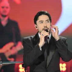 Dawid Kwiatkowski w czarnym garniturze śpiewa do mikrofonu na scenie z muzykiem w tle.