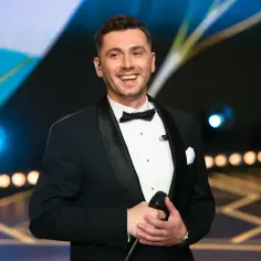 Uśmiechnięty Kamil Bednarek w smokingu, trzymający mikrofon. Myślami pewnie przy swojej żonie.