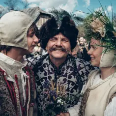 Uśmiechnięta obsada 1670 w strojach z epoki, mężczyzna w futrzanej czapie z pawim piórem, młoda para obok.