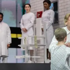 Scena teatralna: grupa osób w białych fartuchach, w tym mężczyzna klęczący przed dzieckiem w piżamie. To może być pierwsza próba, pierwszy cień wątpliwości.