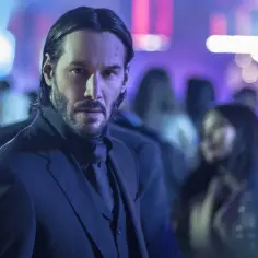Keanu Reeves w garniturze, w tle rozmyte światła i tłum. Ikona kina akcji, znany z wielu filmów.