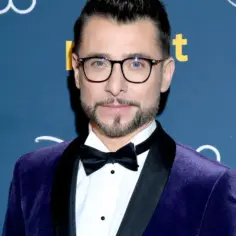 Kacper Kuszewski w fioletowym aksamitnym garniturze z czarną muszką. W tle logo "sat".
