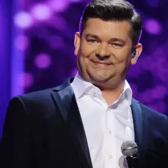 Mężczyzna w czarnym garniturze i białej koszuli, z mikrofonem. Jego niebieskie oczy błyszczą, jakby mówił "akcent przez twe oczy zielone tekst".
