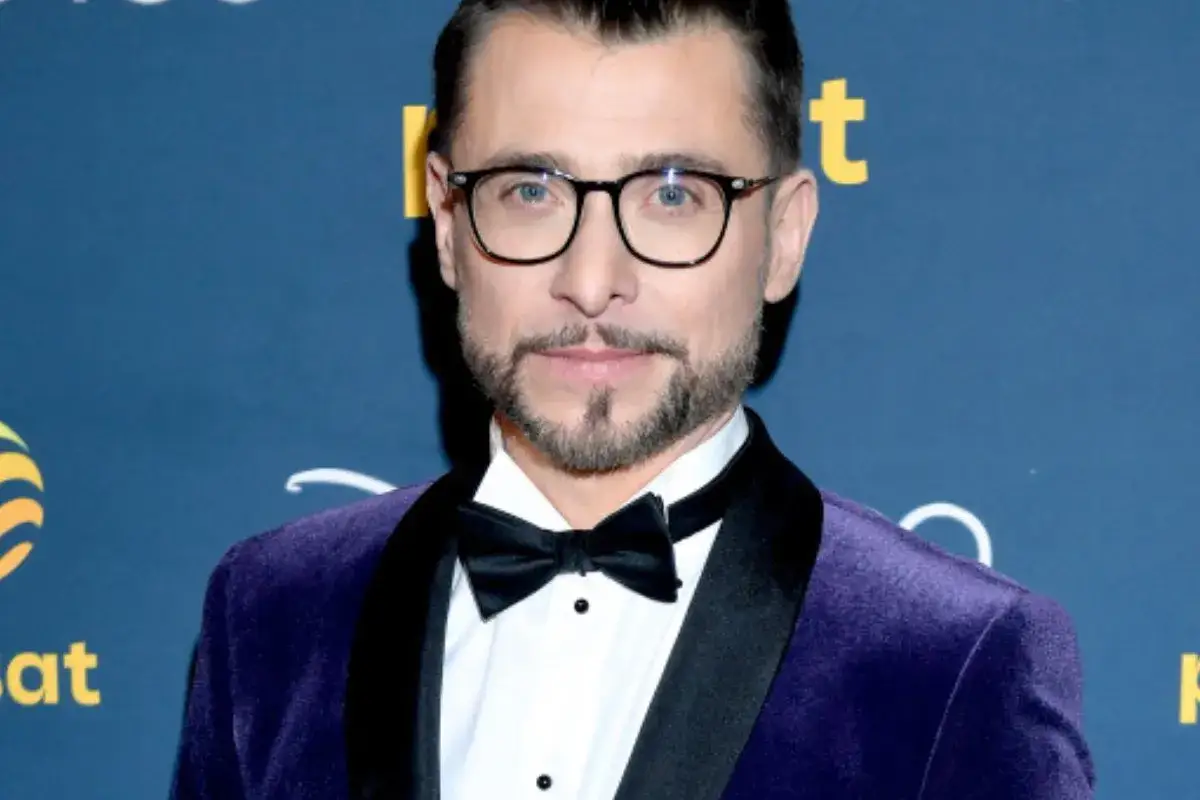 Kacper Kuszewski w fioletowym aksamitnym smokingu z czarną atłasową klapą i czarną muszką. Wygląda elegancko, jakby wybierał się na galę z żoną.