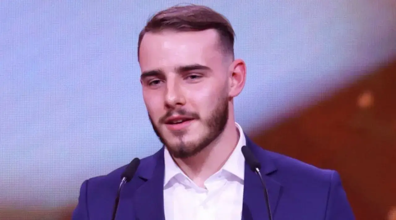 Krystian Ochman, w eleganckim garniturze, przemawia do mikrofonu. Choć na zdjęciu nie widać jego żony, jego uśmiech sugeruje szczęście.