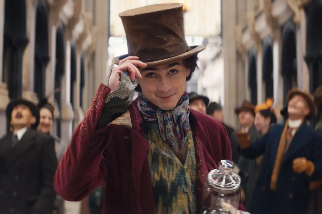 Młody Willy Wonka w kapeluszu i kolorowym stroju, trzymający słoik, zapowiada magiczne przygody w 