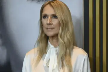 Celine Dion, znana z potężnego głosu, zmaga się z zespołem sztywnego człowieka. Na zdjęciu widać jej blond włosy i białą bluzkę.