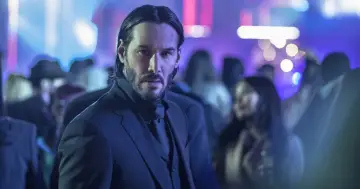 Keanu Reeves w garniturze, w tle rozmyte światła i tłum. Ikona kina akcji, znany z wielu filmów.
