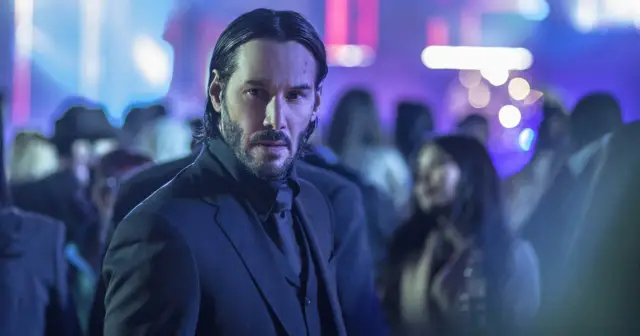 Keanu Reeves w garniturze, w tle rozmyte światła i tłum. Ikona kina akcji, znany z wielu filmów.